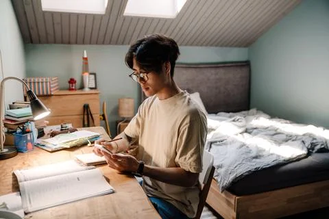 Asian guy using cellphone while sitting at desk and studying at home Stock Photos