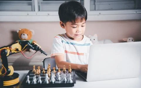 Asian little boy programming code to robot machine arm on laptop for play chess Stock Photos