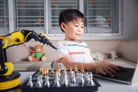 Asian little boy programming code to robot machine arm on laptop for play chess Foto stock