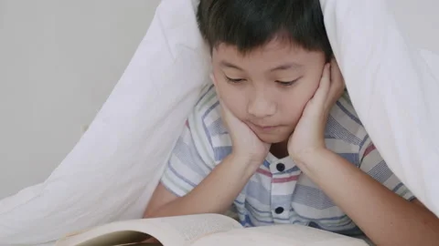 Asian little boy while lying under blanket read book on bedroom at night Stock Footage 164653748
