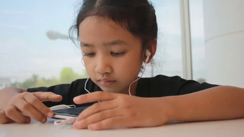 Asian little girl using smartphone with in ear headphone and sitting near gla Stock Footage