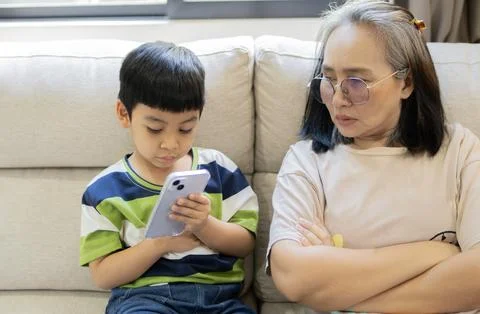 An Asian little son using a smartphone, while frustrated mother is watching.. Stock Photos