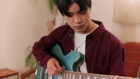 Asian male relaxes and plays his blue guitar on the couch at home. Stock Footage 220757341