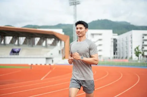 Asian male runner smiling while jogging on running track in sports stadium. Stock Photos