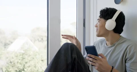 Asian man 20s checking phone wearing white headset gazing out window closing Stock Footage 342112943