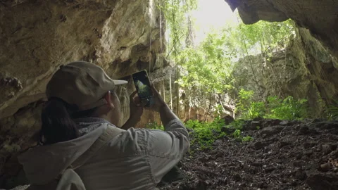Asian man adventurer using a smartphone to take photos towards a cave skylight Stock Footage 335797928