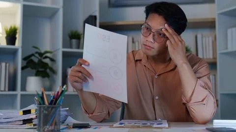 Asian man analyzing financial document with serious expression working late Stockfoto's