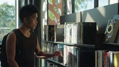 Asian man in backpack browses through books in bright library Stock Footage 87450933