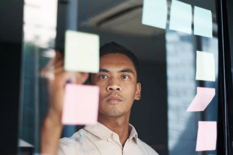 An Asian man brainstorms with sticky notes as seen through a window Foto stock