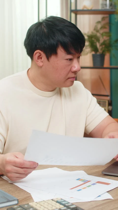Asian man comparing paper reports with laptop data, focused expression reveals Stock Footage 317657405
