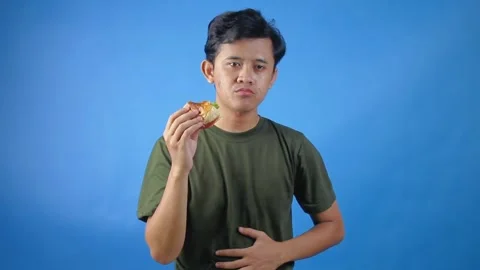 Asian man eating hamburger until full while holding his stomach Stock Footage 323779489