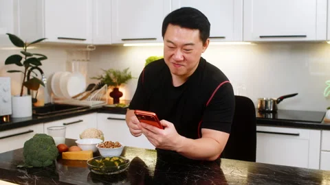 Asian man eats broccoli near smartphone in kitchen while reading health news and Stock Footage 347035366