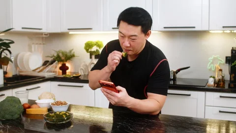 Asian man eats fresh broccoli snack in kitchen while reading nutrition article Stock Footage 347035011