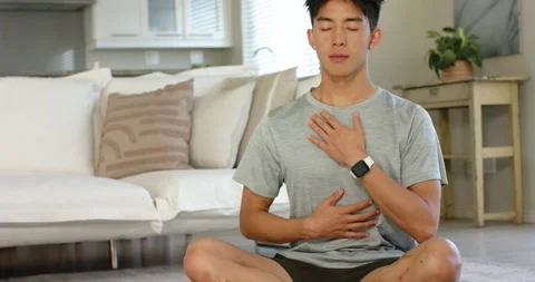 Asian man entering frame sitting on green yoga mat in living room practicing Stock Footage 338325864
