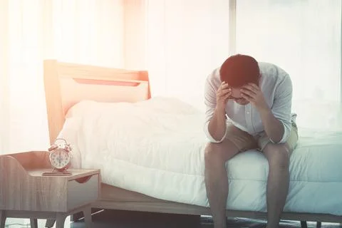Asian Man is having headache while waking up with alarm clock on bed in the m Stock Photos