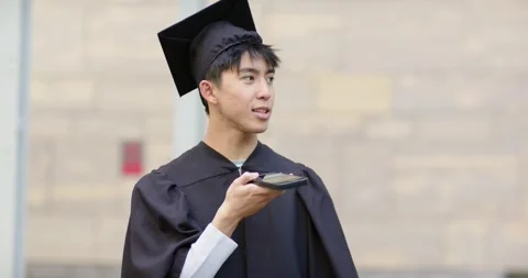 Asian man holding device and wearing cap and gown at campus practicing Stock Footage 341320150