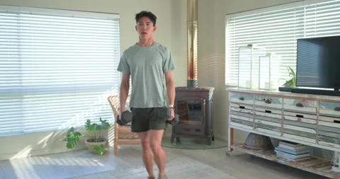 Asian man holding hex weights doing walking lunges, checking watch for reps in Stock Footage 345071311