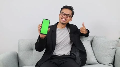 Asian man at home, sitting on the couch and showing the camera a green mock-up Video stock 336509997