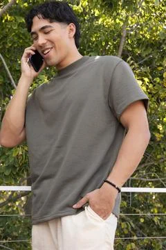 Asian man leaning on cable railing amid leaves wearing olive tee beaded bracelet Stock-Fotos