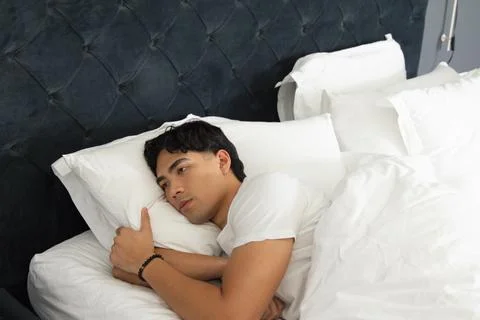 Asian man lying on bed hugging pillow while wearing white T-shirt and beaded Stock Photos