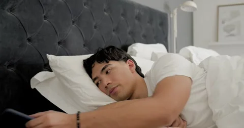 Asian man lying on bed scrolling smartphone eyes growing heavy placing phone Stock Footage 339416273