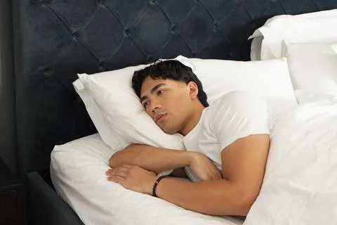 Asian man lying right-side on bed with white bedding, wearing white T-shirt and Stock Photos