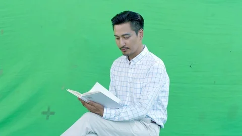 Asian man reading and looking up from book on green background Stock Footage 80806787