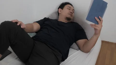 Asian man reading book in bed before sleep, minimalist sparse room with sin.. Video stock 295992862