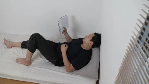 Asian man reading book in bed before sleep, minimalist sparse room with sin.. Video stock 295992963