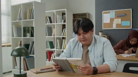 Asian Man Reading Book in Library with Diverse Classmates Stock Footage 337485160