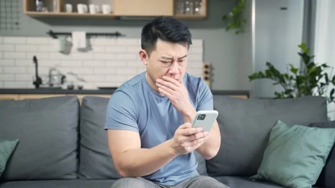 Asian man reading sms bad news on smartphone phone sitting on sofa at home.  Stock Footage 191194802