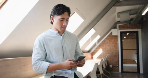 Asian man receiving smartphone notification scrolling on bench in loft glancing Stock Footage 339958983