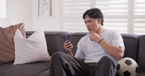 Asian man scrolling smartphone on couch with soccer ball, reading confusing text Stock Footage 339321184
