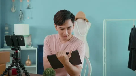 An Asian man is showing clothes in front of smartphone  live streaming at his Vídeos de archivo 172007834