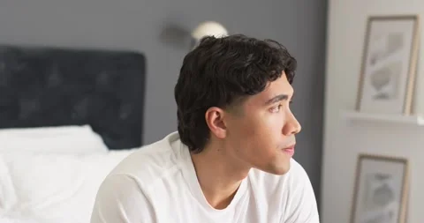 Asian man sitting on bed edge in bedroom, turning toward framed prints, wearing Stock Footage 336175317