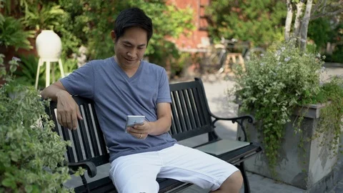 Asian man sitting on a bench using smartphone in garden. Stock Footage 126505847