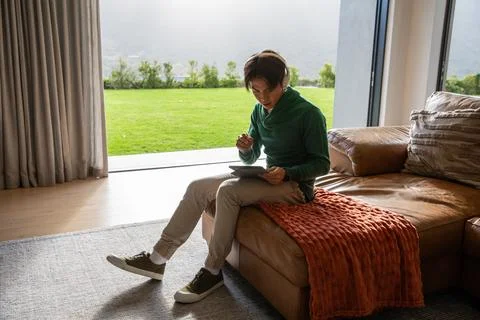 Asian man sitting on brown leather sofa using tablet with stylus in modern Stock Photos