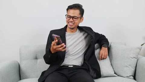 Asian man sitting on the couch and showing the camera a green mock-up of a Video stock 336510068