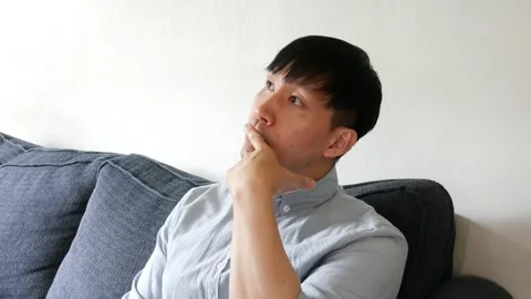 Asian man sitting on a couch looking up at something and nodding Stock Footage 247480020