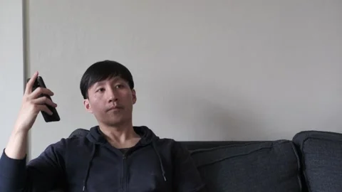 Asian man sitting on a couch using his cell phone Stock Footage 303256437
