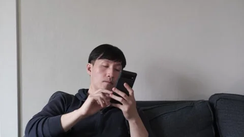 Asian man sitting on a couch using his cell phone Stock Footage 303256438