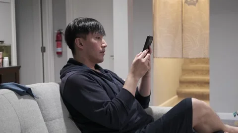 Asian man sitting on a couch using his cell phone Stock Footage 303256485