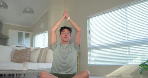 Asian man sitting on green mat in living room, breathing, lifting arms, joining Stock Footage 337705176