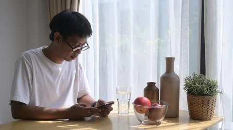 Asian man sitting at kitchen table and using smart phone for checking news or 스톡 사진