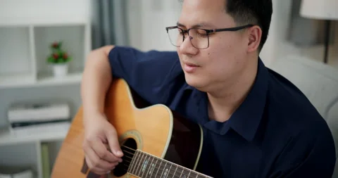 Asian man sitting on sofa playing acoustic guitar in living room at home. Stock-Footage 305723909