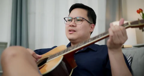 Asian man sitting on sofa playing acoustic guitar in living room at home. Stock-Footage 305724393
