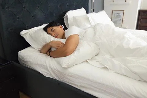 Asian man sleeping on bed in white T-shirt, hugging pillow near tufted headboard Stock Photos