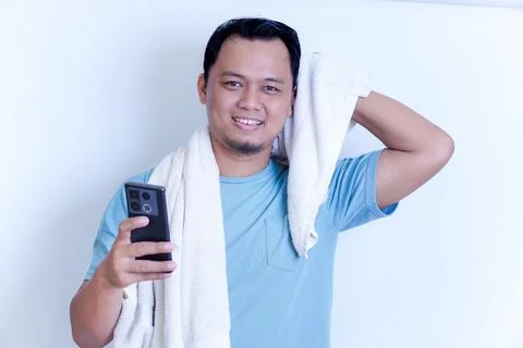 A Asian man is smiling while looking at his phone, holding a towel over his.. Stock Photos