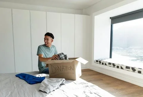 Asian man smiling while packing folded clothing into labeled DONATION box on bed Photos