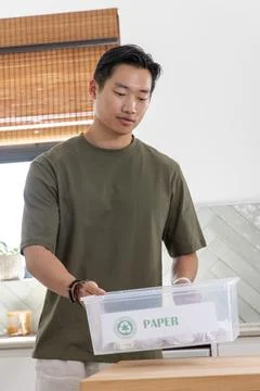 Asian man standing at kitchen counter lifting clear recycling bin labeled PAPER, Stock Photos